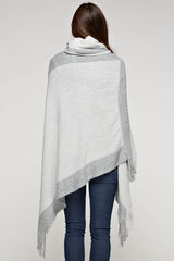 [Color: Grey/Gunmetal] A brunette model wearing a two-tone border knit poncho with Lurex accents at the border and fringe trim. Featuring a cozy cowl neckline and asymmetrical hem, this poncho offers a perfect mix of style and comfort.