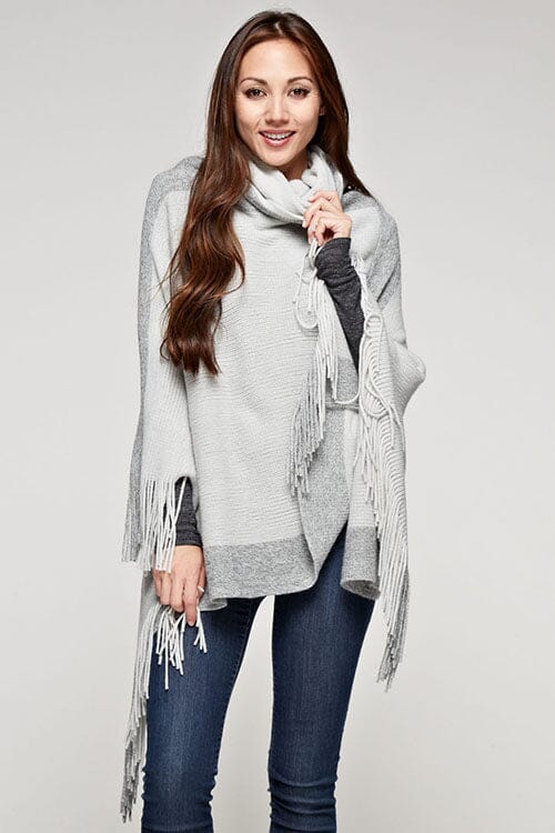 [Color: Grey/Gunmetal] A brunette model wearing a two-tone border knit poncho with Lurex accents at the border and fringe trim. Featuring a cozy cowl neckline and asymmetrical hem, this poncho offers a perfect mix of style and comfort.