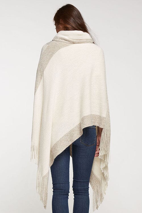 [Color: Copper] A brunette model wearing a two-tone border knit poncho with Lurex accents at the border and fringe trim. Featuring a cozy cowl neckline and asymmetrical hem, this poncho offers a perfect mix of style and comfort.