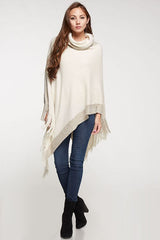 [Color: Copper] A brunette model wearing a two-tone border knit poncho with Lurex accents at the border and fringe trim. Featuring a cozy cowl neckline and asymmetrical hem, this poncho offers a perfect mix of style and comfort.