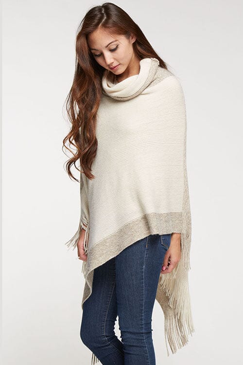 [Color: Copper] A brunette model wearing a two-tone border knit poncho with Lurex accents at the border and fringe trim. Featuring a cozy cowl neckline and asymmetrical hem, this poncho offers a perfect mix of style and comfort.