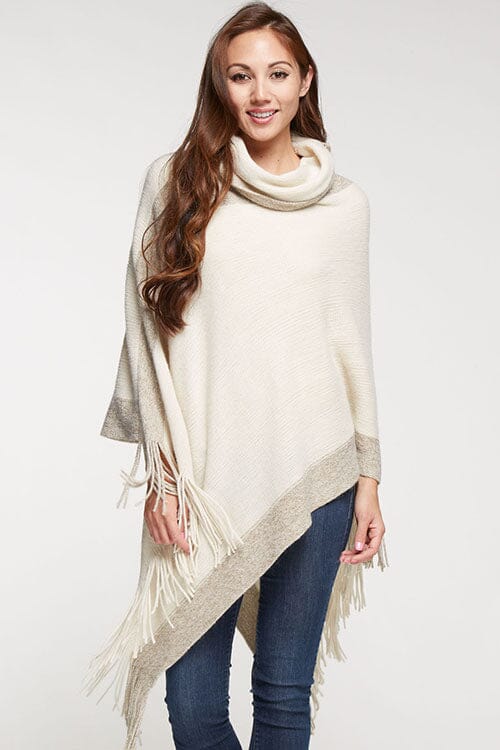 [Color: Copper] A brunette model wearing a two-tone border knit poncho with Lurex accents at the border and fringe trim. Featuring a cozy cowl neckline and asymmetrical hem, this poncho offers a perfect mix of style and comfort.