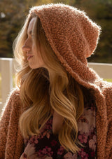 [Color: Camel] A detail  image of a blonde model wearing a fuzzy brown fall cardigan with a popcorn texture at a farm. The cardigan features an open front, large cozy patch pockets, and an attached hoodie, draped for warmth and ideal for layering.