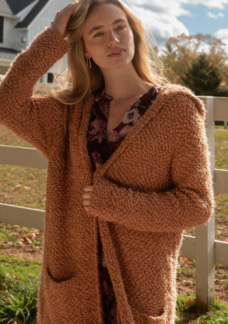 [Color: Camel] A front facing image of a blonde model wearing a fuzzy brown fall cardigan with a popcorn texture at a farm. The cardigan features an open front, large cozy patch pockets, and an attached hoodie, draped for warmth and ideal for layering.