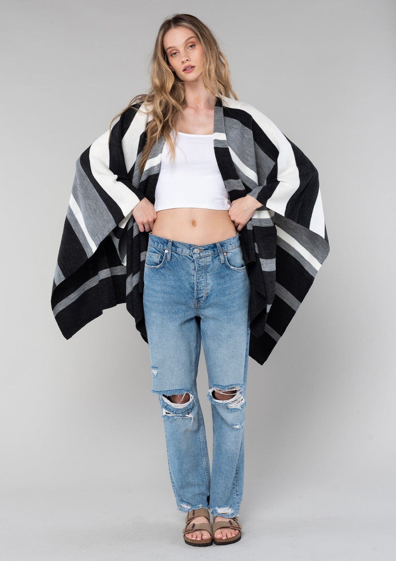 [Color: Charcoal Combo] A blonde model wearing a charcoal combo multi-stripe poncho with an open front, asymmetrical hemline, and a casual, effortless drape in soft acrylic knit.