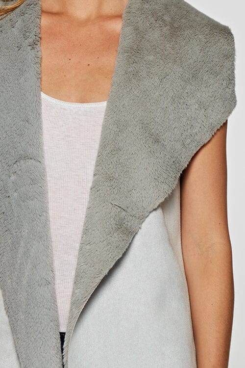 [Color: Grey] A blonde model wearing a casual grey vegan suede vest with a faux fur trim interior and contrasting ribbed knit back. 