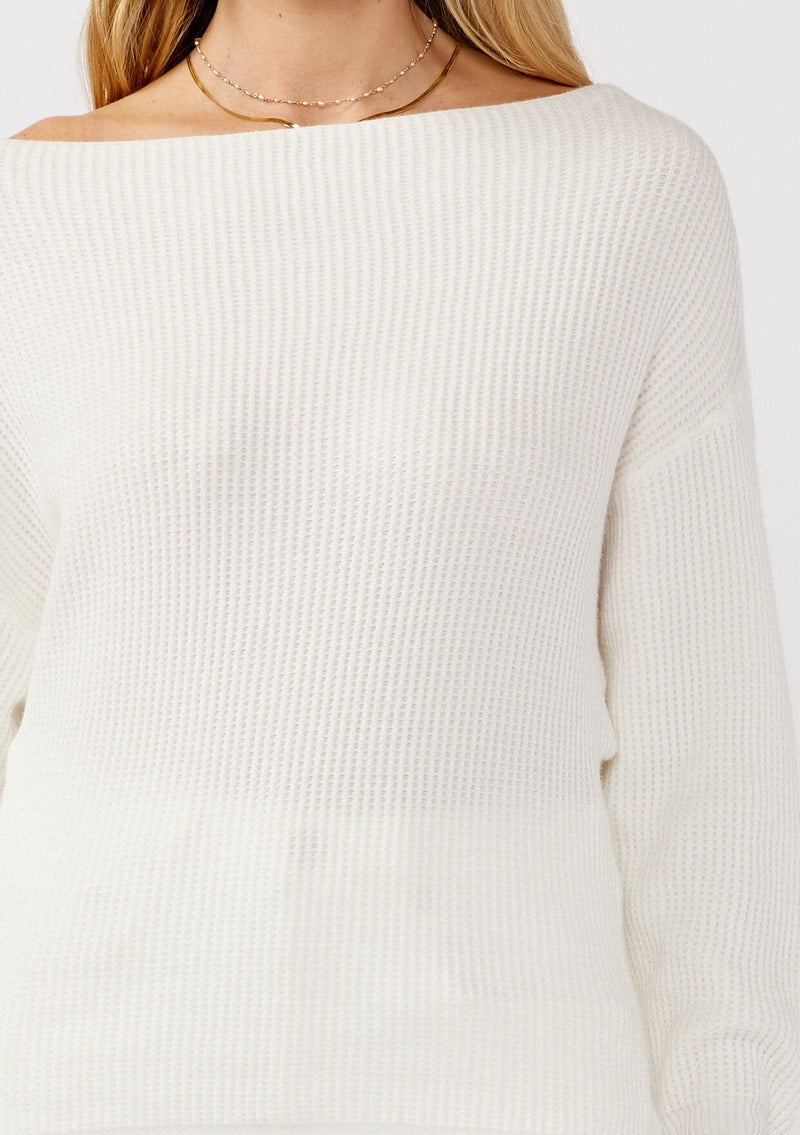 [Color: Winter White] A detail image of a blonde model wearing a white waffle knit pullover sweater. With long sleeves, a relaxed fit, and a wide neckline that can be worn off the shoulder.