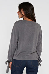 [Color: Heather Charcoal] A brunette model wearing a grey pullover sweater with a long sleeves, dropped shoulders, and a tie wrist detail with grommets. 