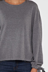 [Color: Heather Charcoal] A brunette model wearing a grey pullover sweater with a long sleeves, dropped shoulders, and a tie wrist detail with grommets. 