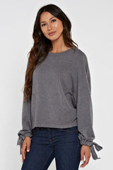 [Color: Heather Charcoal] A brunette model wearing a grey pullover sweater with a long sleeves, dropped shoulders, and a tie wrist detail with grommets. 