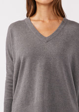 [Color: Heather Charcoal] A detailed image of a brunette model wearing a soft and relaxed fit knit sweater in gray. A cozy fall pullover sweater with a boxy silhouette, v neckline, ribbed trim, and side vents. 