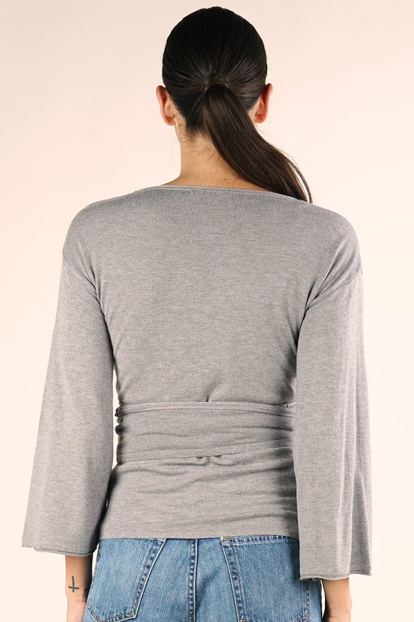 [Color: Heather Ash] A brunette model wearing a casual grey wrap front top with long flowy sleeves, a v neckline, and an adjustable waist tie. 