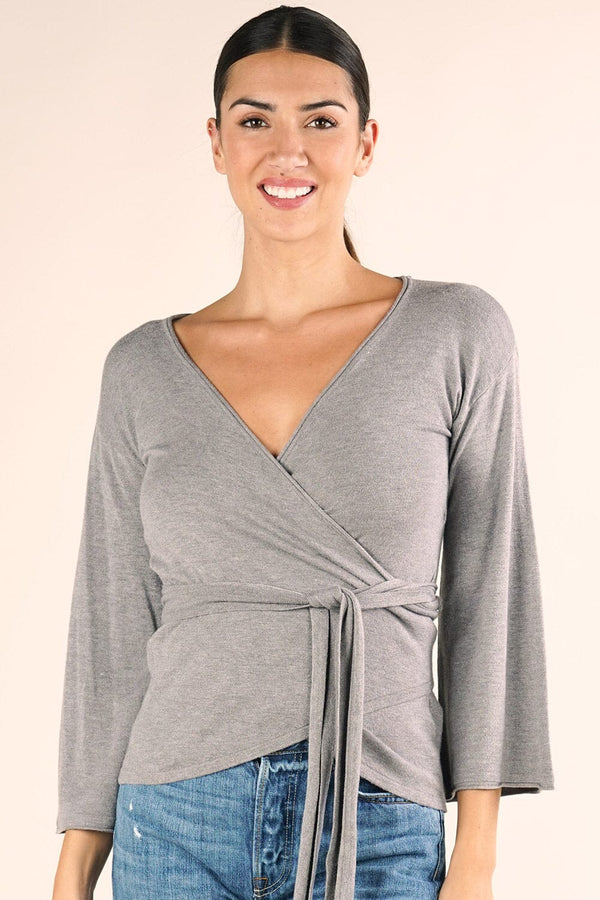 [Color: Heather Ash] A brunette model wearing a casual grey wrap front top with long flowy sleeves, a v neckline, and an adjustable waist tie. 