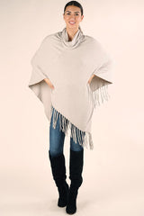 [Color: Heather Stone] A front facing image of a brunette model wearing a light beige cable knit rib sweater poncho. With a cowl neckline and an asymmetric fringed hemline.