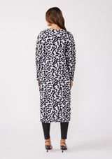 [Color: Snow Leopard] A brunette model wearing an ultra soft and fuzzy statement cardigan in a bold allover snow leopard print. A duster length sweater featuring oversize front patch pockets. Styled here with black skinny jeans and heels for a formal yet casual look. 