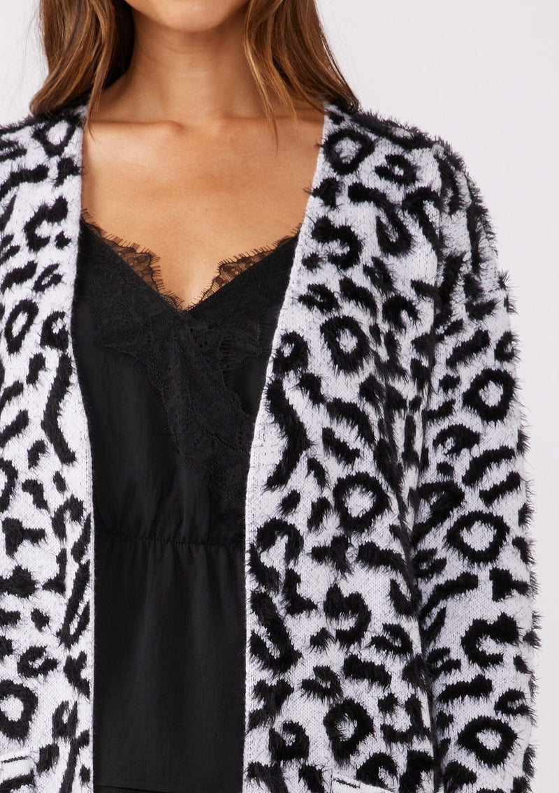 [Color: Snow Leopard] A brunette model wearing an ultra soft and fuzzy statement cardigan in a bold allover snow leopard print. A duster length sweater featuring oversize front patch pockets. 