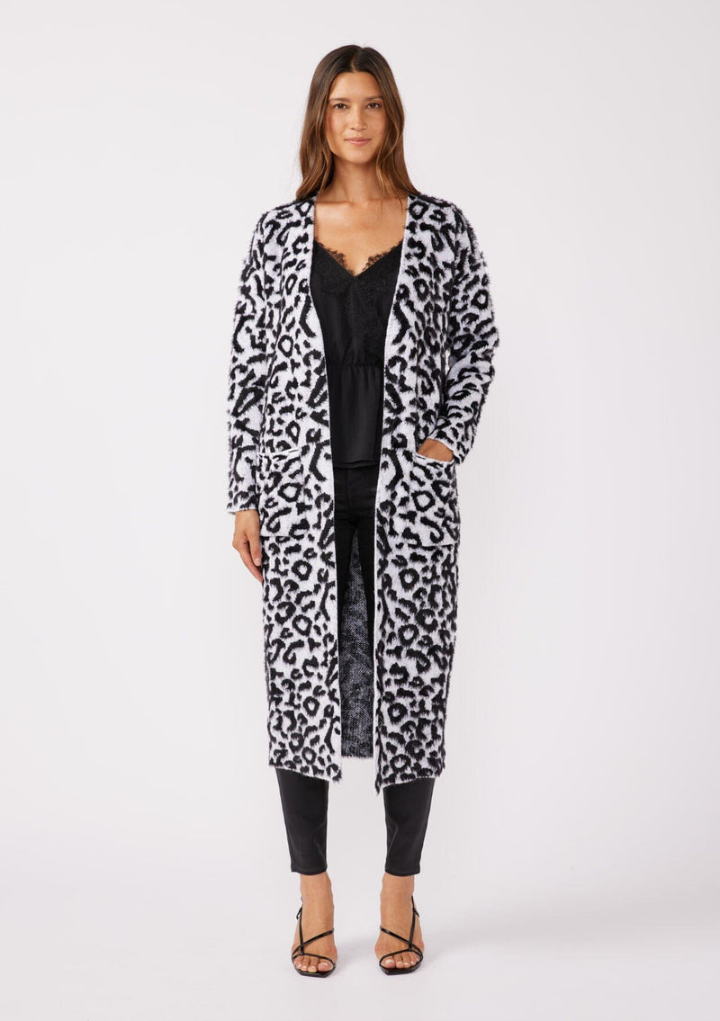 [Color: Snow Leopard] A brunette model wearing an ultra soft and fuzzy statement cardigan in a bold allover snow leopard print. A duster length sweater featuring oversize front patch pockets. Styled here with black skinny jeans and heels for a formal yet casual look. 