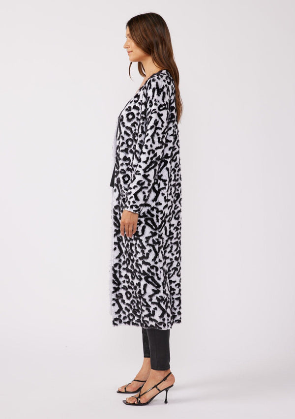 [Color: Snow Leopard] A brunette model wearing an ultra soft and fuzzy statement cardigan in a bold allover snow leopard print. A duster length sweater featuring oversize front patch pockets. Styled here with black skinny jeans and heels for a formal yet casual look. 