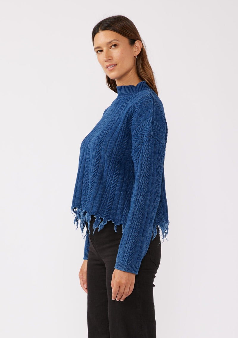 [Color: Indigo] A relaxed fit indigo blue cable knit sweater. A 100% cotton pullover featuring a fringed hemline, a mock neckline, and a back vent detail. Styled with dark denim for the Fall season. 