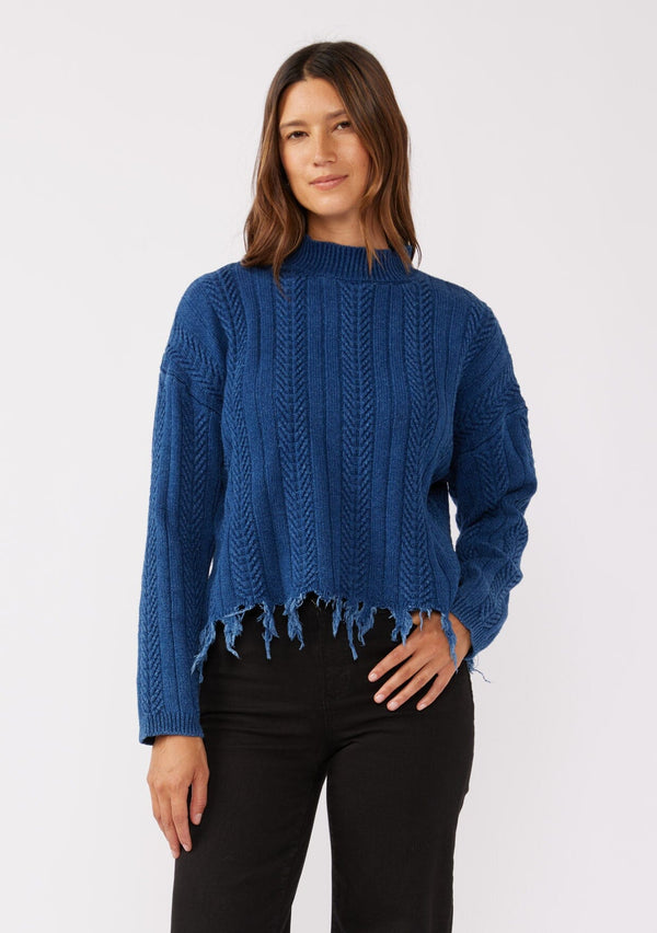 [Color: Indigo] A relaxed fit indigo blue cable knit sweater. A 100% cotton pullover featuring a fringed hemline, a mock neckline, and a back vent detail. Styled with dark denim for the Fall season. 