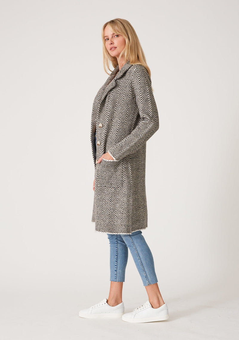 [Color: Taupe/Cream] A side facing image of a blonde model wearing a soft sweater coat in a taupe and cream herringbone pattern. With long sleeves, a button front, side pockets, a mid length hemline, and a notched collar. 