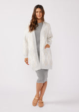 [Color: Heather Grey/Ivory] An image of a brunette model wearing an ultra-soft mid length cardigan in a light grey and ivory argyle plaid pattern. With long sleeves, an open front, and side pockets. Styled with a grey midi dress for a formal yet casual look. 