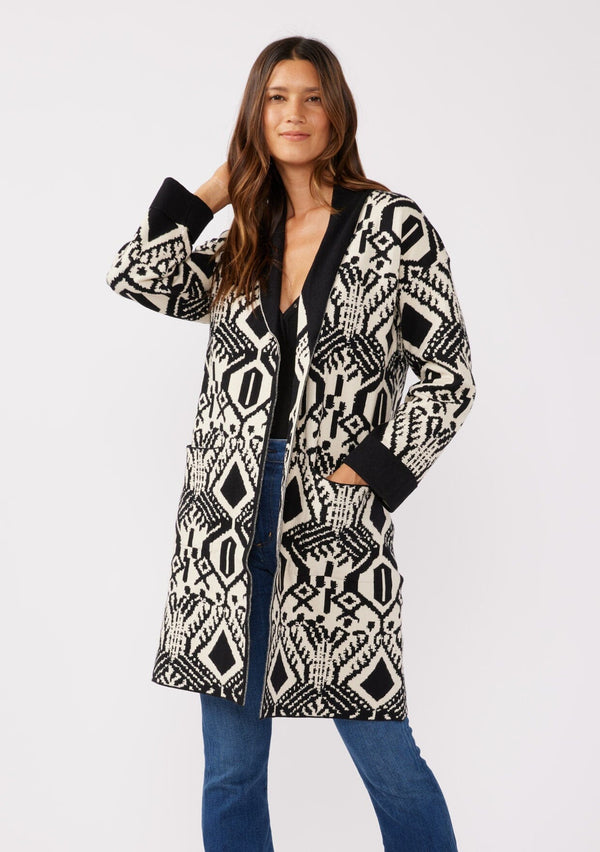 [Color: Natural/Black] A front facing image of a brunette model wearing a natural off white and black bohemian patterned mid length cardigan. A long sweater style with long sleeves, contrast black wrist cuffs, a contrast black shawl collar, an open front, and side pockets.