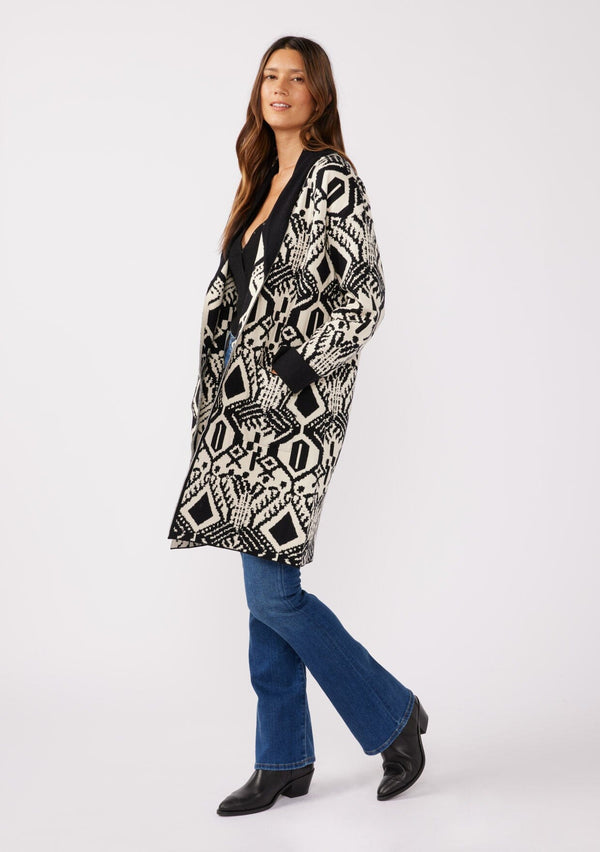 [Color: Natural/Black] A side facing image of a brunette model wearing a natural off white and black bohemian patterned mid length cardigan. A long sweater style with long sleeves, contrast black wrist cuffs, a contrast black shawl collar, an open front, and side pockets.