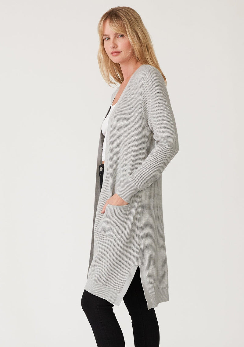 [Color: Heather Silver Grey] A side facing image of a blonde model wearing a light heather grey waffle knit cardigan. A lightweight mid length cardigan sweater with long sleeves, side pockets, and an open front. 