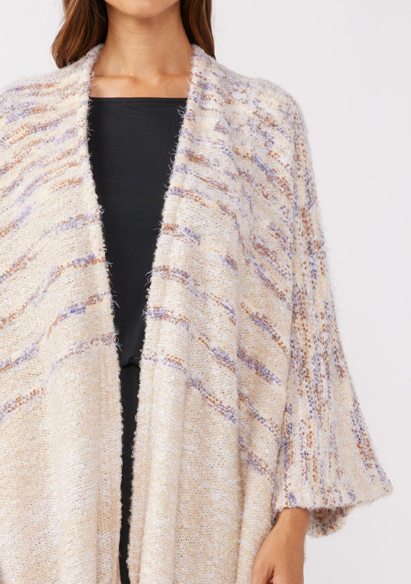 [Color: Oatmeal Multi] A detailed  image of a brunette model wearing a fuzzy bohemian sweater poncho in a cream multi color knit. With an open front and tassel fringe trim. The perfect on the go style when an extra cozy layer is needed. Styled with a casual black maxi for the Fall season. 