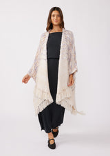 [Color: Oatmeal Multi] A full body front facing image of a brunette model wearing a fuzzy bohemian sweater poncho in a cream multi color knit. With an open front and tassel fringe trim. The perfect on the go style when an extra cozy layer is needed. Styled with a casual black maxi for the Fall season. 