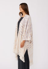 [Color: Oatmeal Multi] A side facing image of a brunette model wearing a fuzzy bohemian sweater poncho in a cream multi color knit. With an open front and tassel fringe trim. The perfect on the go style when an extra cozy layer is needed. Styled with a casual black maxi for the Fall season. 