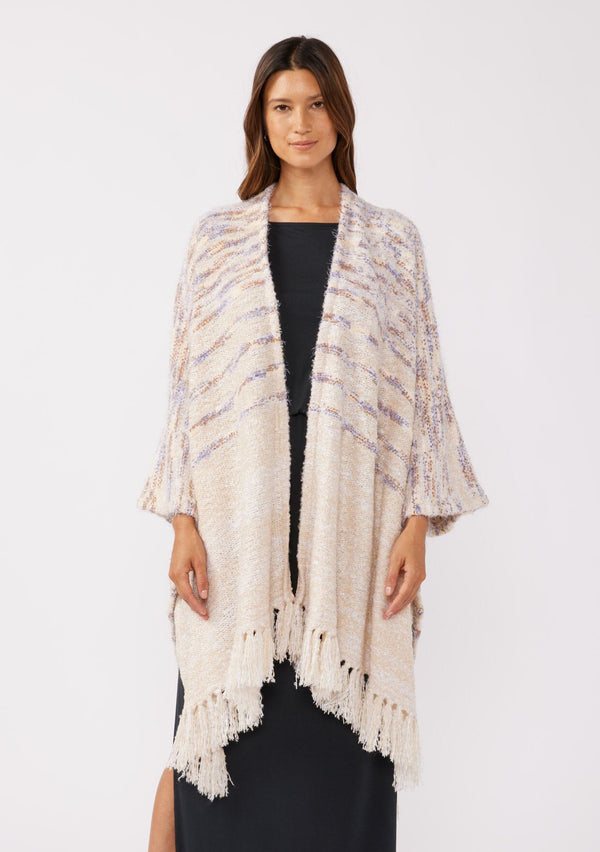 [Color: Oatmeal Multi] A front facing image of a brunette model wearing a fuzzy bohemian sweater poncho in a cream multi color knit. With an open front and tassel fringe trim. The perfect on the go style when an extra cozy layer is needed. Styled with a casual black maxi for the Fall season. 