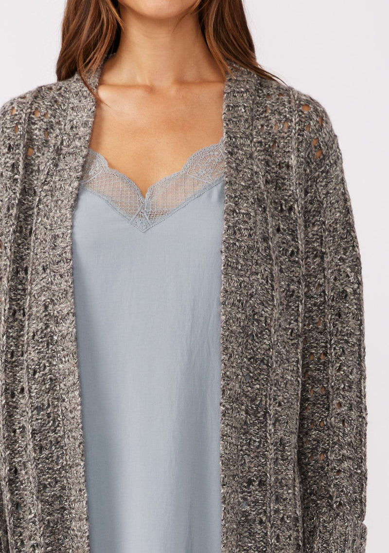 [Color: Charcoal] A detailed image of a brunette model wearing a dark charcoal grey open knit cardigan sweater. A mid length lightweight cardigan with long sleeves, an open front, and ribbed wrist cuffs.