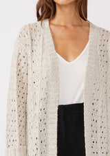 [Color: Oatmeal] A detailed image of a brunette model wearing an oatmeal open knit cardigan sweater. A mid length lightweight cardigan with long sleeves, an open front, and ribbed wrist cuffs.