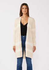 [Color: Eggshell] A front facing image of a brunette model wearing a cream colored fuzzy bohemian mid length cardigan in a textured patchwork knit. With long sleeves and an open front.