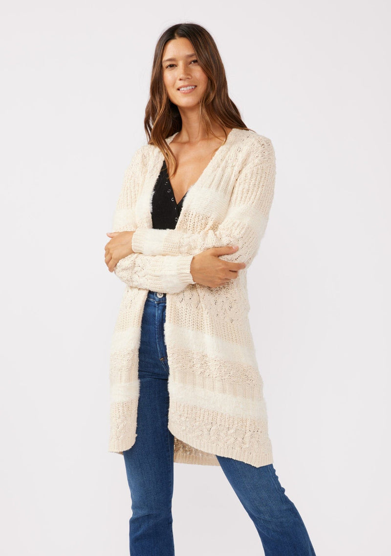 [Color: Eggshell] A front facing image of a brunette model wearing a cream colored fuzzy bohemian mid length cardigan in a textured patchwork knit. With long sleeves and an open front.