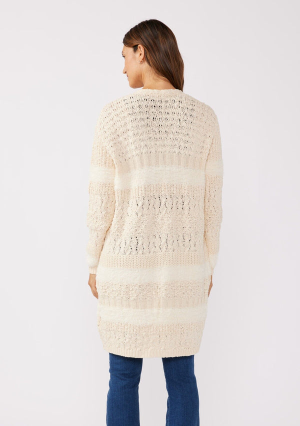 [Color: Eggshell] A back facing image of a brunette model wearing a cream colored fuzzy bohemian mid length cardigan in a textured patchwork knit. With long sleeves and an open front.