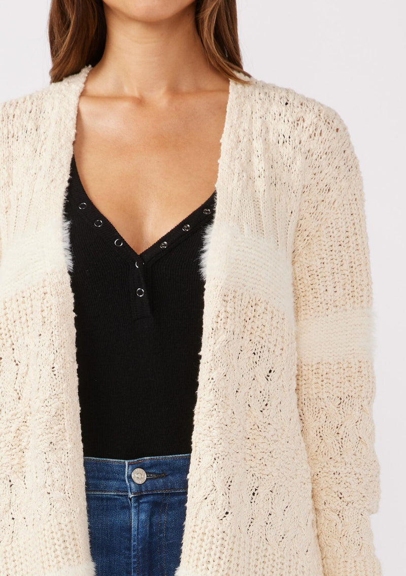 [Color: Eggshell] A detailed image of a brunette model wearing a cream colored fuzzy bohemian mid length cardigan in a textured patchwork knit. With long sleeves and an open front.