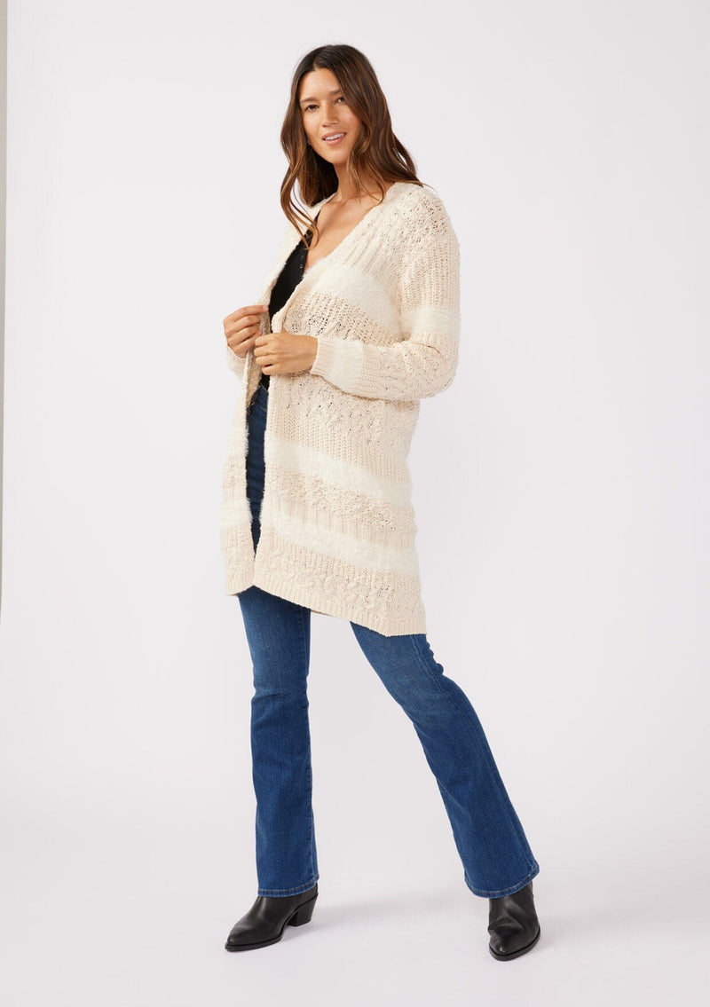 [Color: Eggshell] A side facing image of a brunette model wearing a cream colored fuzzy bohemian mid length cardigan in a textured patchwork knit. With long sleeves and an open front.