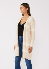 [Color: Eggshell] A side facing image of a brunette model wearing a cream colored fuzzy bohemian mid length cardigan in a textured patchwork knit. With long sleeves and an open front.