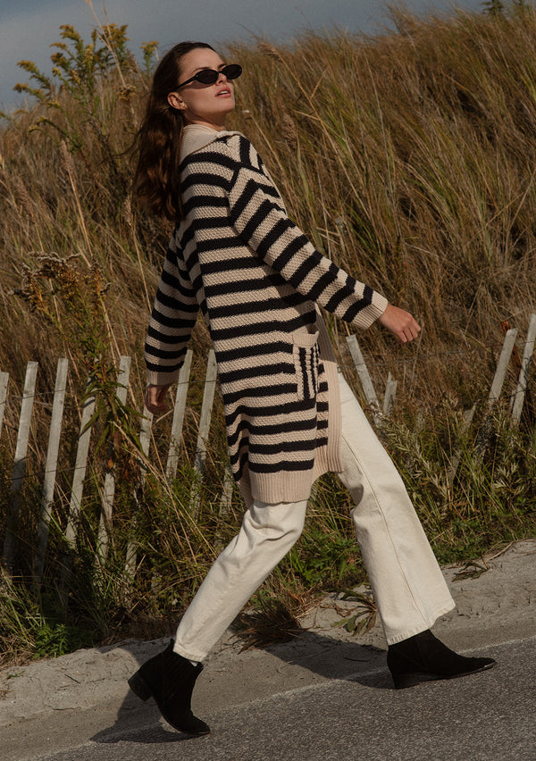 [Color: Navy/Natural] A side facing image of a brunette model wearing a mid length cardigan in a black and white stripe. With long sleeves, a collared neckline, an open front, and side pockets.