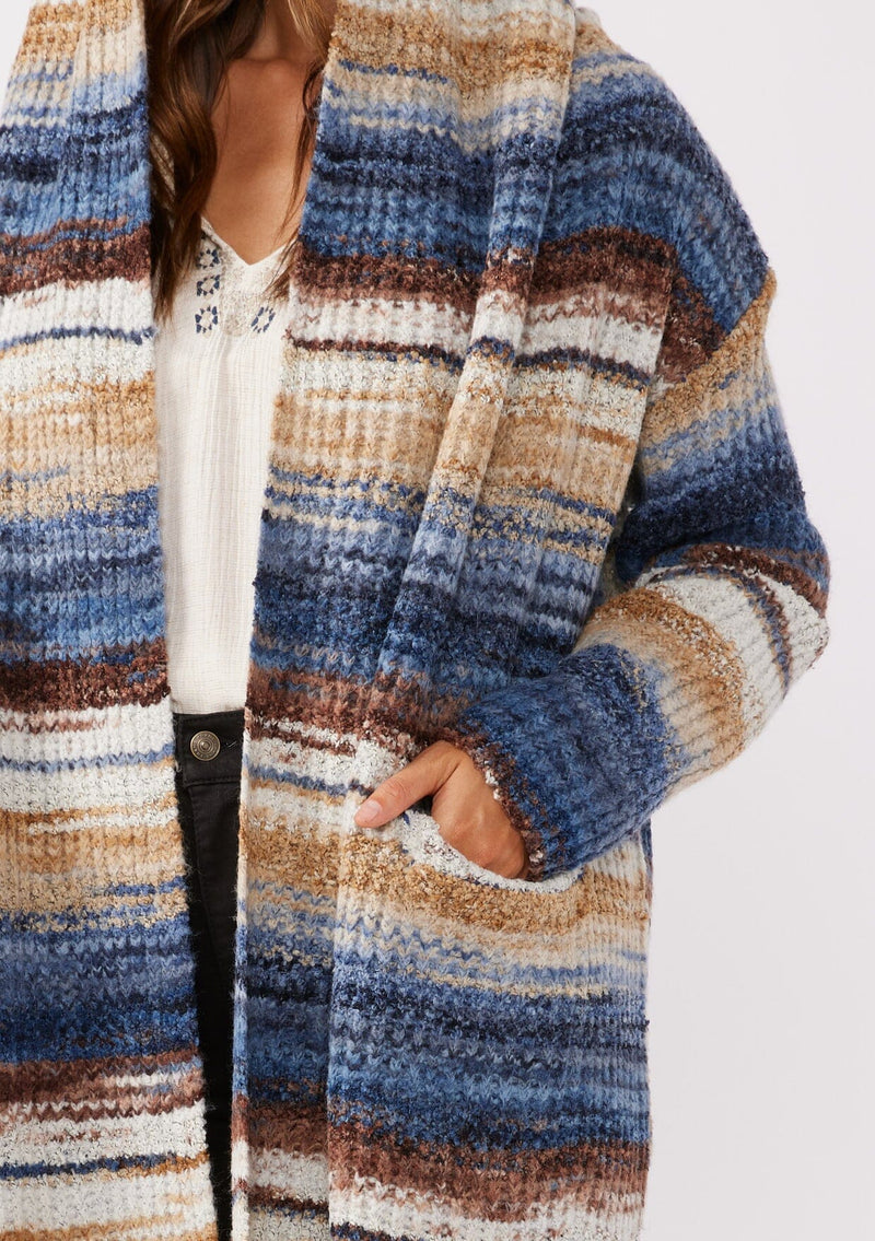 [Color: Navy/Taupe] A detailed image of a brunette model wearing a cozy bohemian space dye chunky knit cardigan in a navy blue multi-color stripe pattern. This plush cardigan features long sleeves, a cozy hood, essential pockets, and an open front. The long length and thick knit make it the perfect sweater for the fall season.