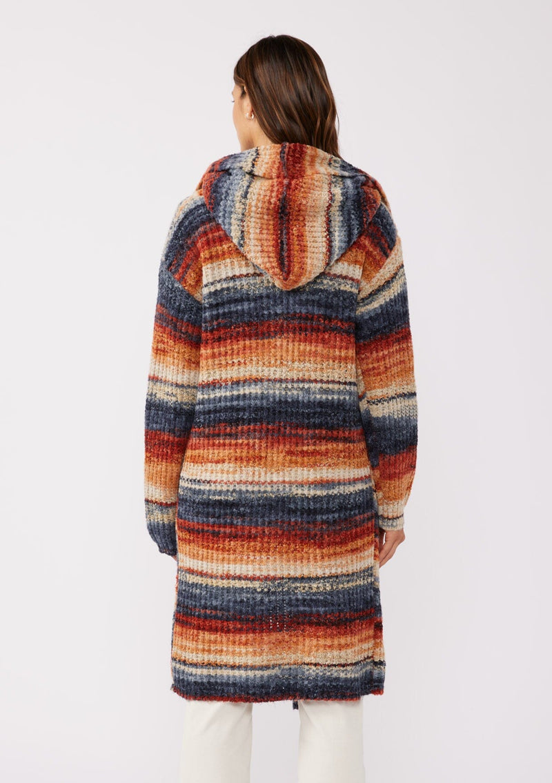 [Color: Rust/Navy] A back facing image with a brunette model wearing a cozy bohemian space dye chunky knit cardigan in a rust multi-color stripe pattern. This plush cardigan features long sleeves, a cozy hood, essential pockets, and an open front. The long length and thick knit make it the perfect sweater for the fall season.