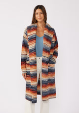 [Color: Rust/Navy] A front facing image with a brunette model wearing a cozy bohemian space dye chunky knit cardigan in a rust multi-color stripe pattern. This plush cardigan features long sleeves, a cozy hood, essential pockets, and an open front. The long length and thick knit make it the perfect sweater for the fall season.