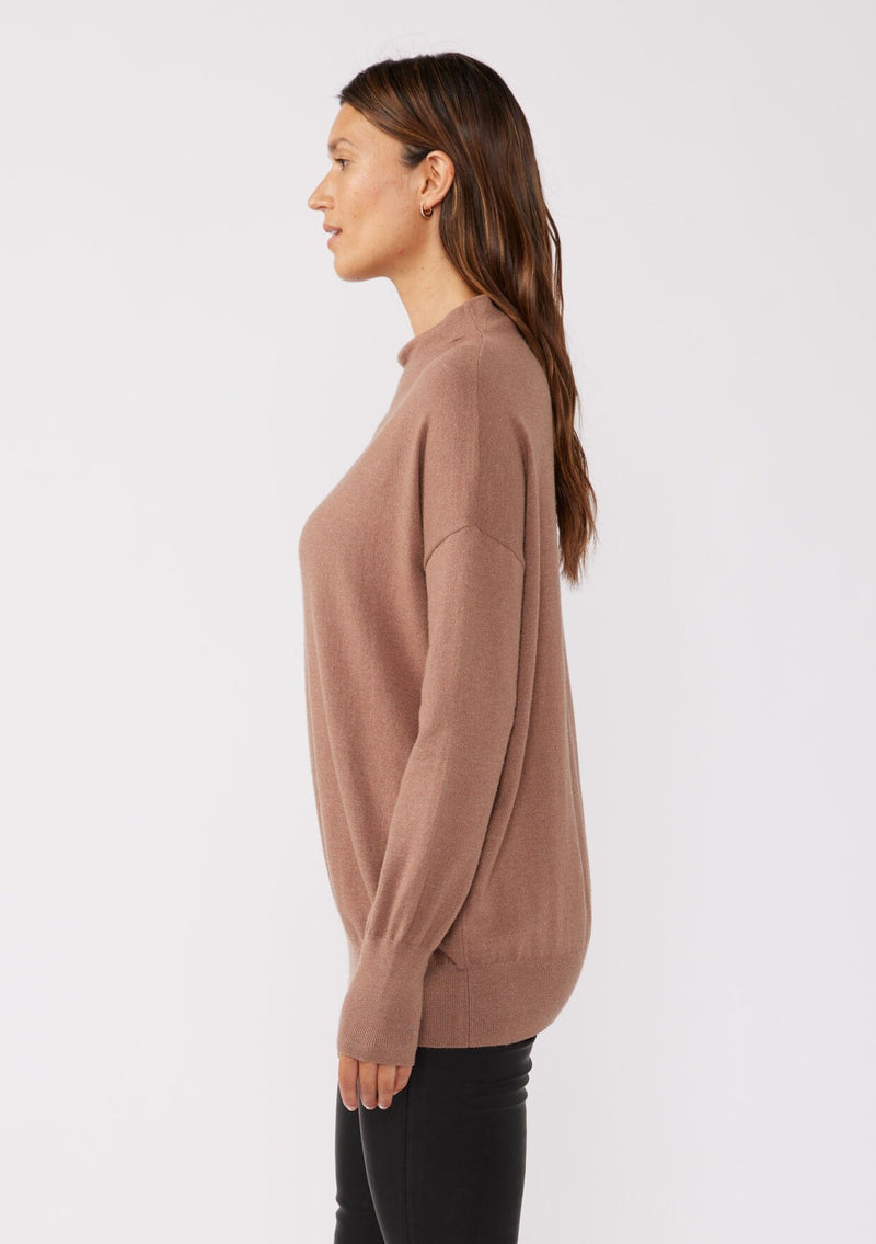 [Color: Chestnut] A side facing image of a brunette model wearing a brown cozy lightweight pullover with a mock neckline. Complete with long sleeves, dropped shoulders, and a snugged ribbed trim. Perfect for layering or styling on its own for a relaxed, laid-back fall look. 