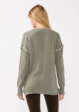 [Color: Sage/Natural] A blonde model wearing an everyday green sweater with a relaxed silhouette. With a classic v neckline, long sleeves, and an exposed seam with blanket stitch. A fall sweater styled with black bottom and casual sandals. 