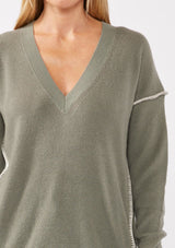 [Color: Sage/Natural] A blonde model wearing an everyday green sweater with a relaxed silhouette. With a classic v neckline, long sleeves, and an exposed seam with blanket stitch. A fall sweater styled with black bottom and casual sandals. 