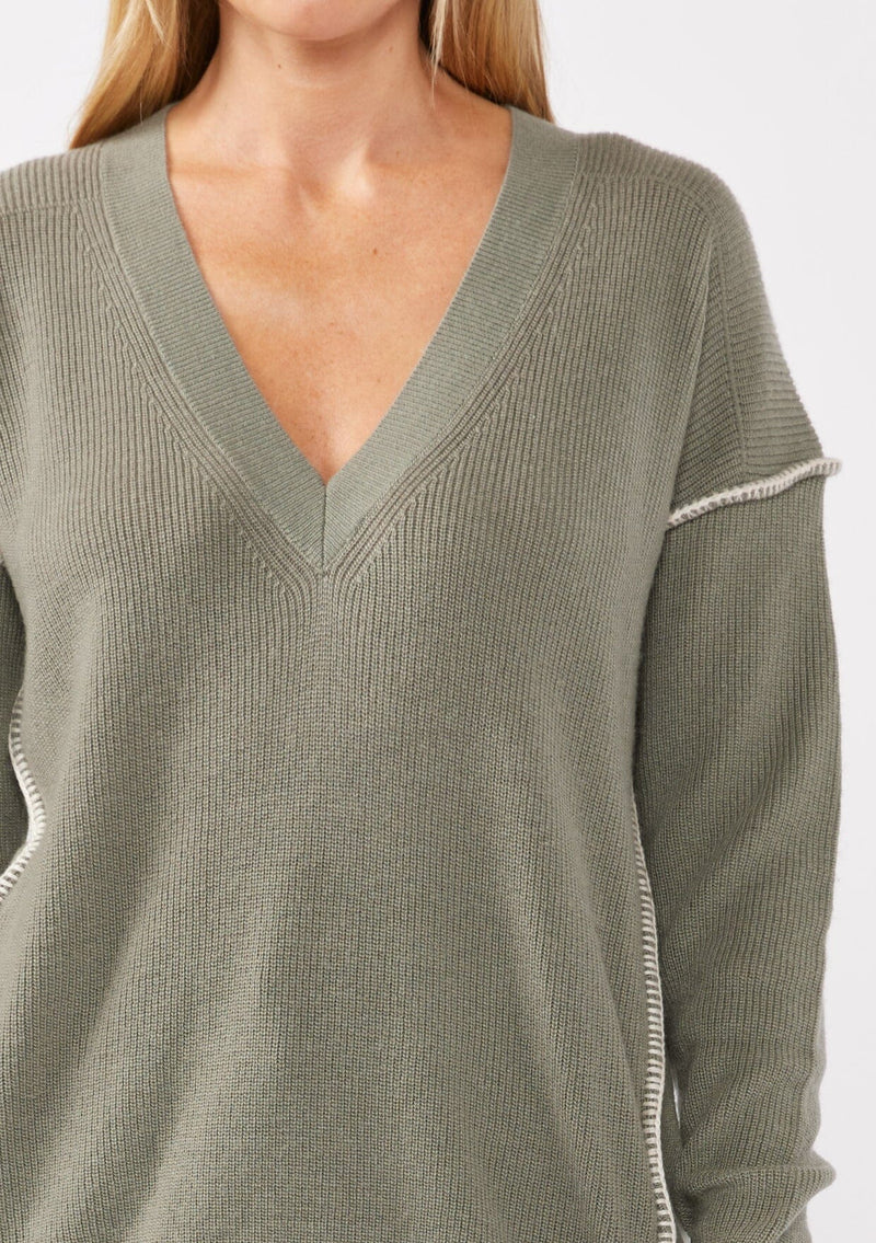 [Color: Sage/Natural] A blonde model wearing an everyday green sweater with a relaxed silhouette. With a classic v neckline, long sleeves, and an exposed seam with blanket stitch. A fall sweater styled with black bottom and casual sandals. 