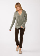 [Color: Sage/Natural] A blonde model wearing an everyday green sweater with a relaxed silhouette. With a classic v neckline, long sleeves, and an exposed seam with blanket stitch. A fall sweater styled with black bottom and casual sandals. 